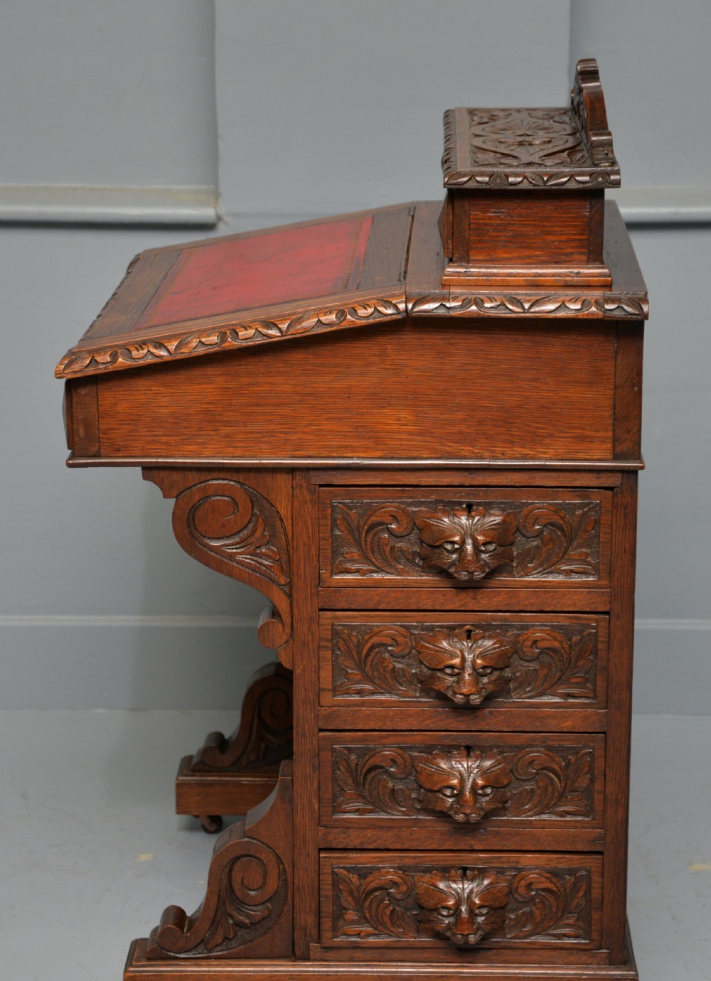 A Victorian dark carved oak Davenport desk featuring a red leather writing slant, carved Green Man mask handles on four side drawers, and a raised stationery gallery. View XVI