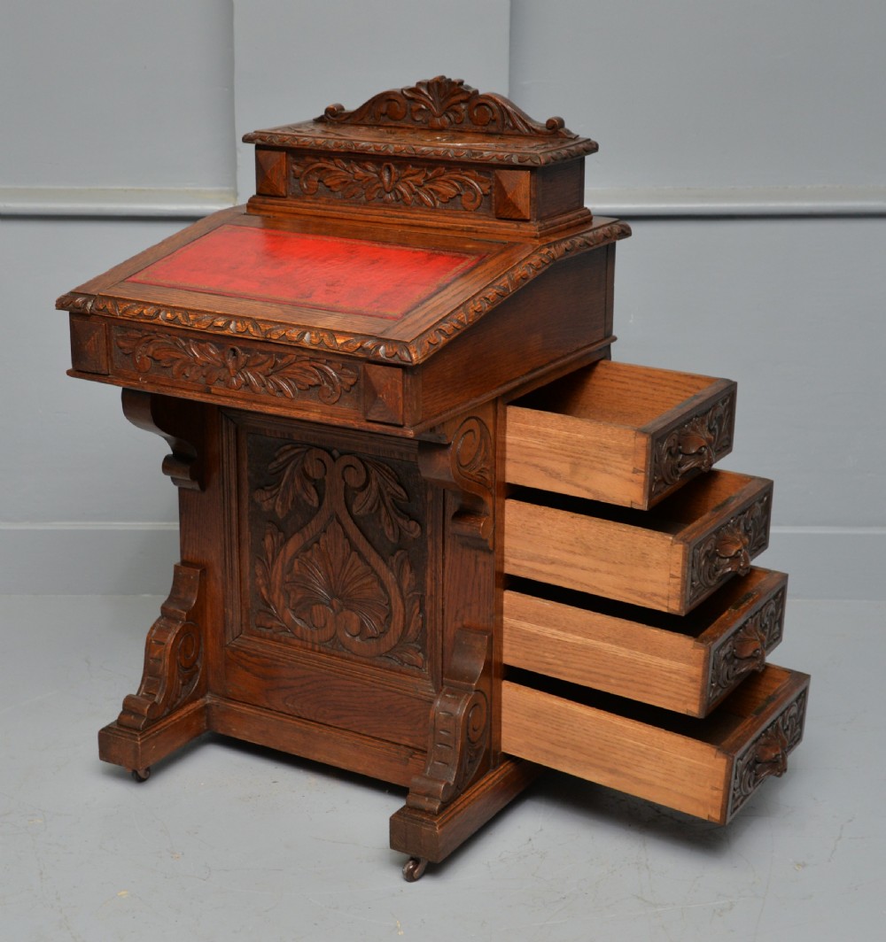 A Victorian dark carved oak Davenport desk featuring a red leather writing slant, carved Green Man mask handles on four side drawers, and a raised stationery gallery. View XX