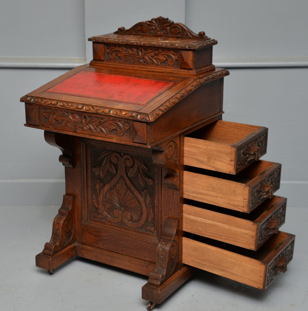 A Victorian dark carved oak Davenport desk featuring a red leather writing slant, carved Green Man mask handles on four side drawers, and a raised stationery gallery. View XXI