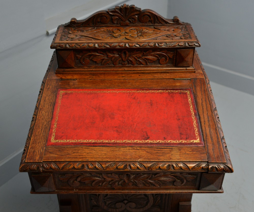 A Victorian dark carved oak Davenport desk featuring a red leather writing slant, carved Green Man mask handles on four side drawers, and a raised stationery gallery. View XXIII