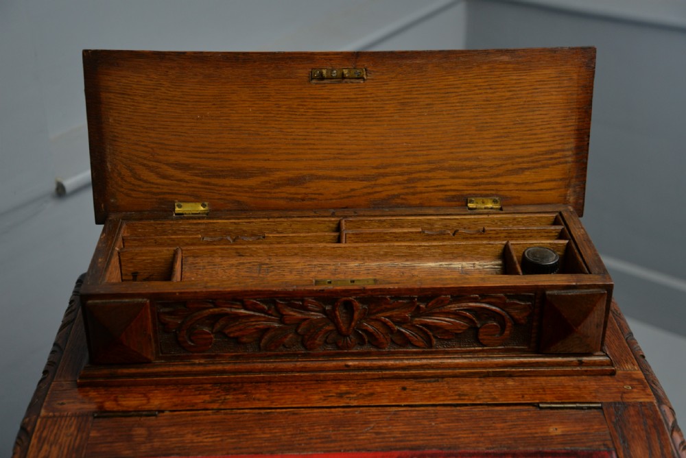 A Victorian dark carved oak Davenport desk featuring a red leather writing slant, carved Green Man mask handles on four side drawers, and a raised stationery gallery. View XXIV