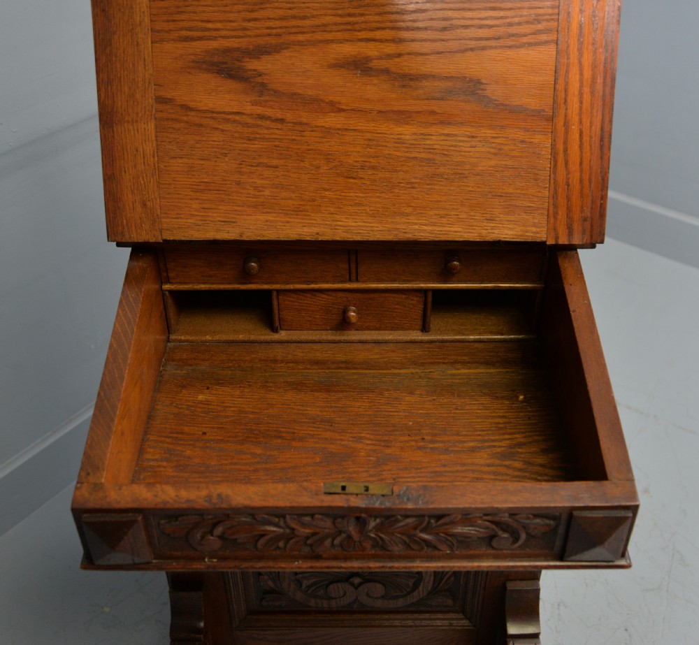 A Victorian dark carved oak Davenport desk featuring a red leather writing slant, carved Green Man mask handles on four side drawers, and a raised stationery gallery. View XXV