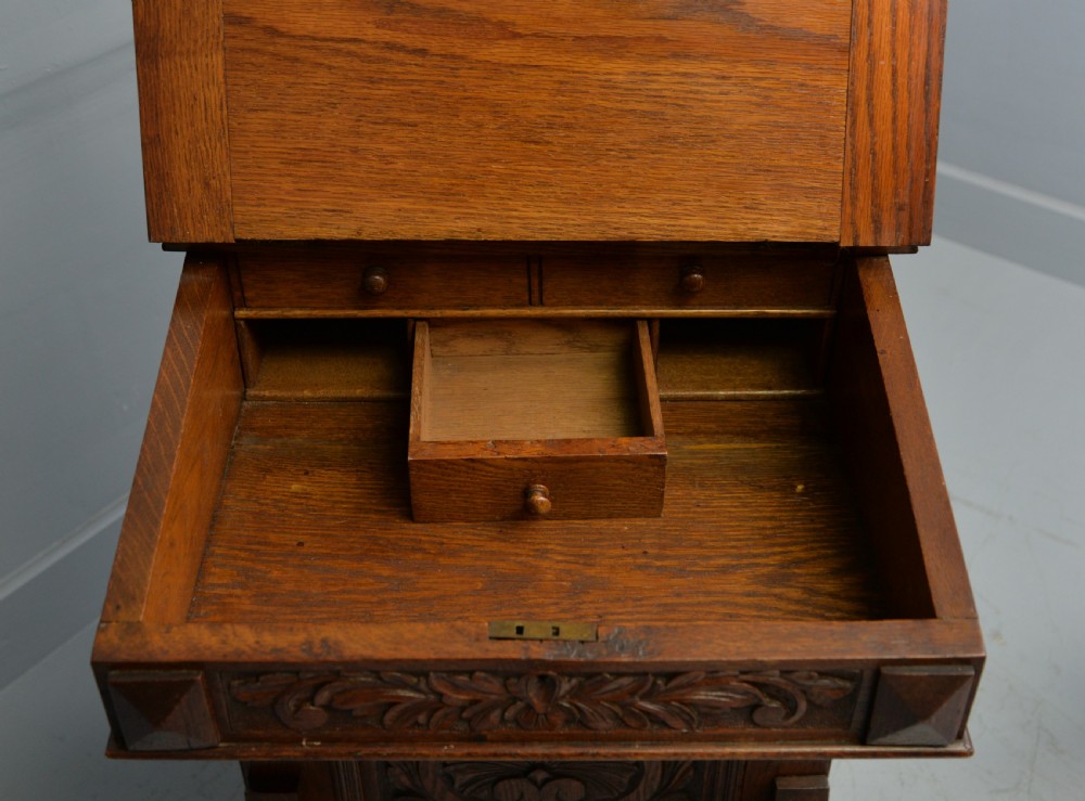 A Victorian dark carved oak Davenport desk featuring a red leather writing slant, carved Green Man mask handles on four side drawers, and a raised stationery gallery. View XXVI
