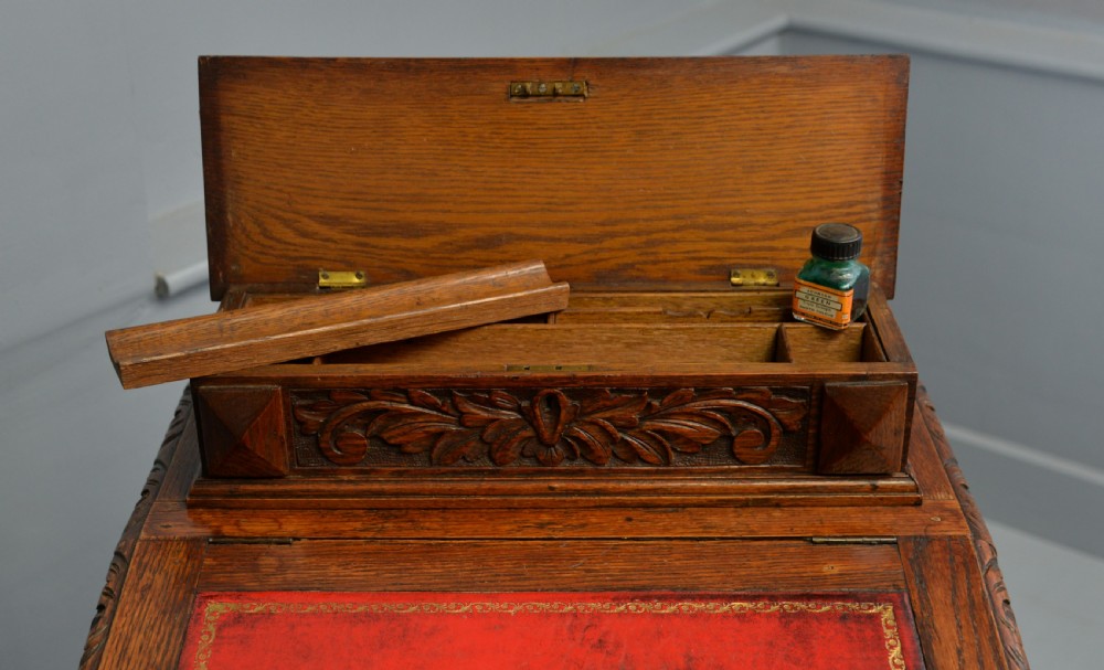 A Victorian dark carved oak Davenport desk featuring a red leather writing slant, carved Green Man mask handles on four side drawers, and a raised stationery gallery. View XXVII