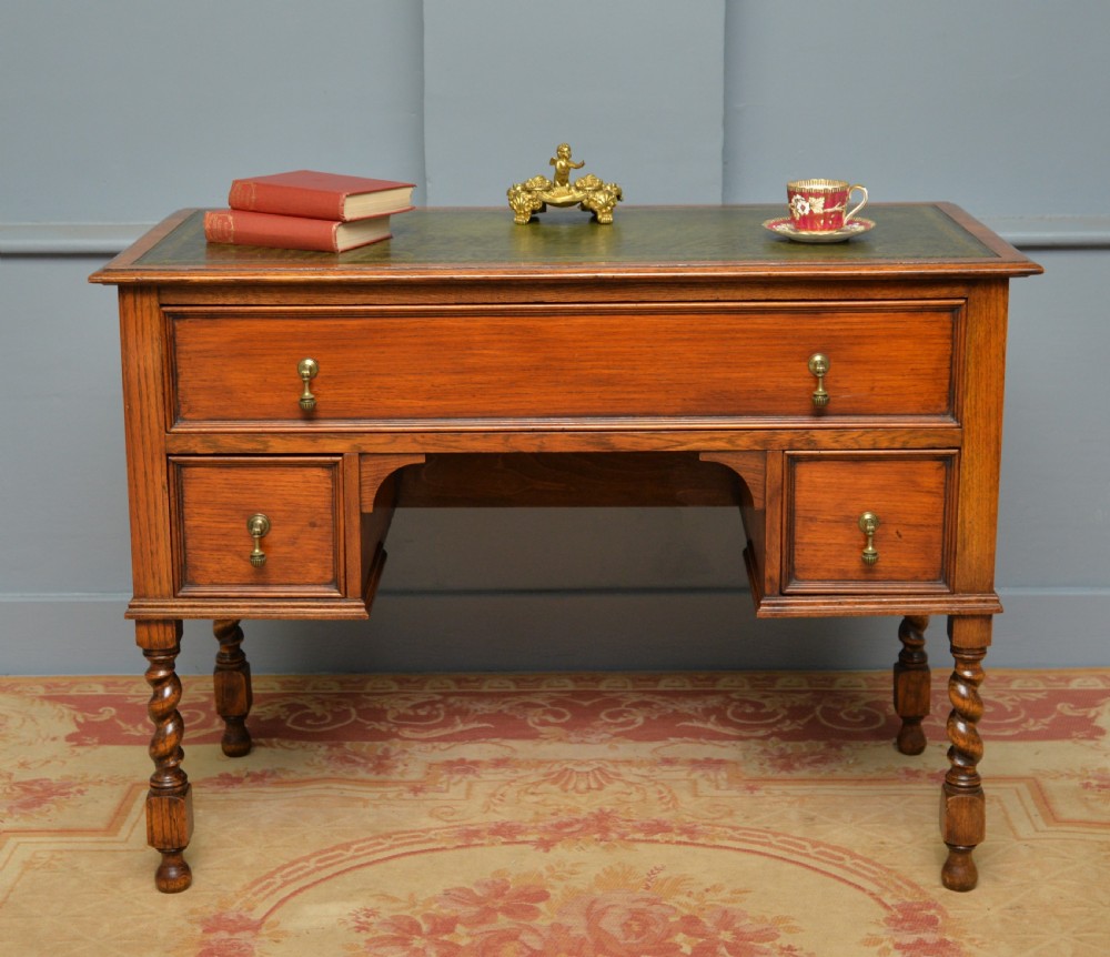 A solid oak writing table featuring a green leather inset top, three drawers with brass drop handles, and turned barley twist legs. View II