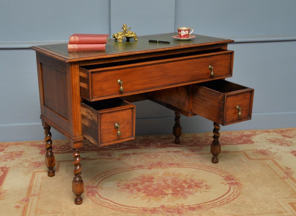 A solid oak writing table featuring a green leather inset top, three drawers with brass drop handles, and turned barley twist legs. View VI