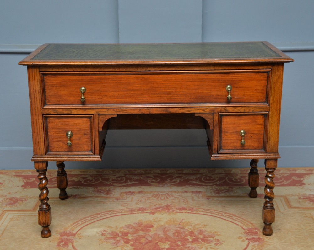 A solid oak writing table featuring a green leather inset top, three drawers with brass drop handles, and turned barley twist legs. View VII