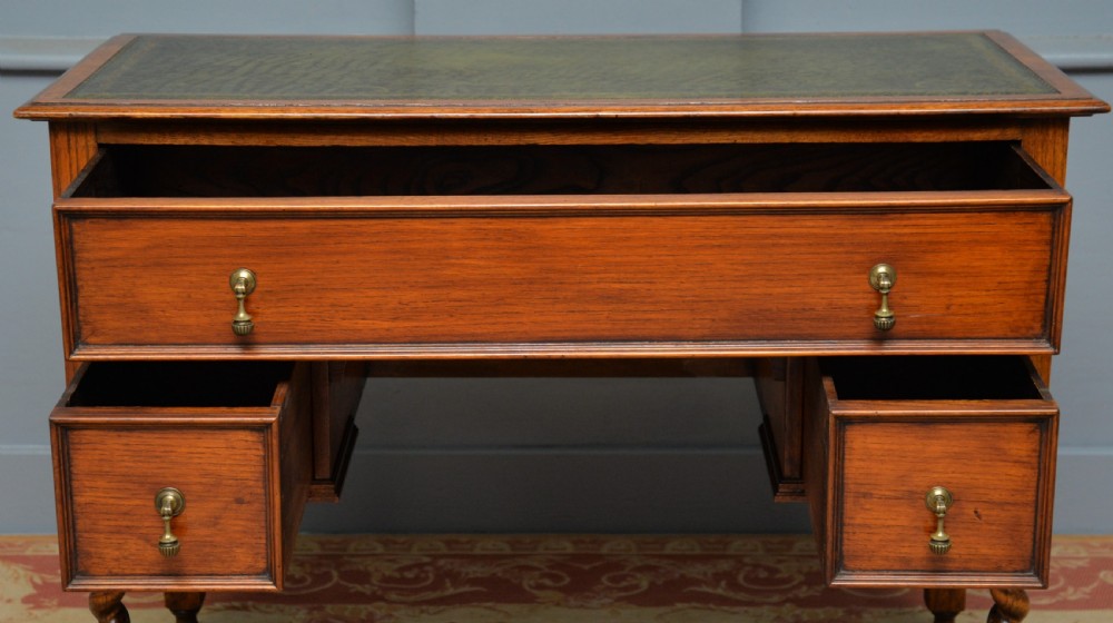 A solid oak writing table featuring a green leather inset top, three drawers with brass drop handles, and turned barley twist legs. View IX