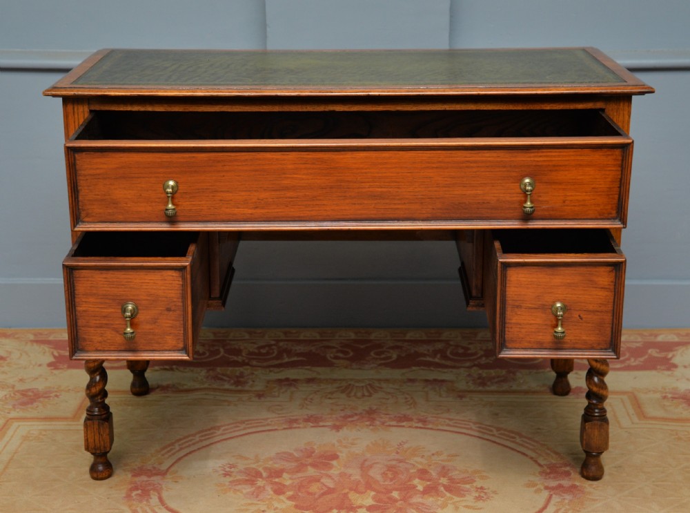 A solid oak writing table featuring a green leather inset top, three drawers with brass drop handles, and turned barley twist legs. View X