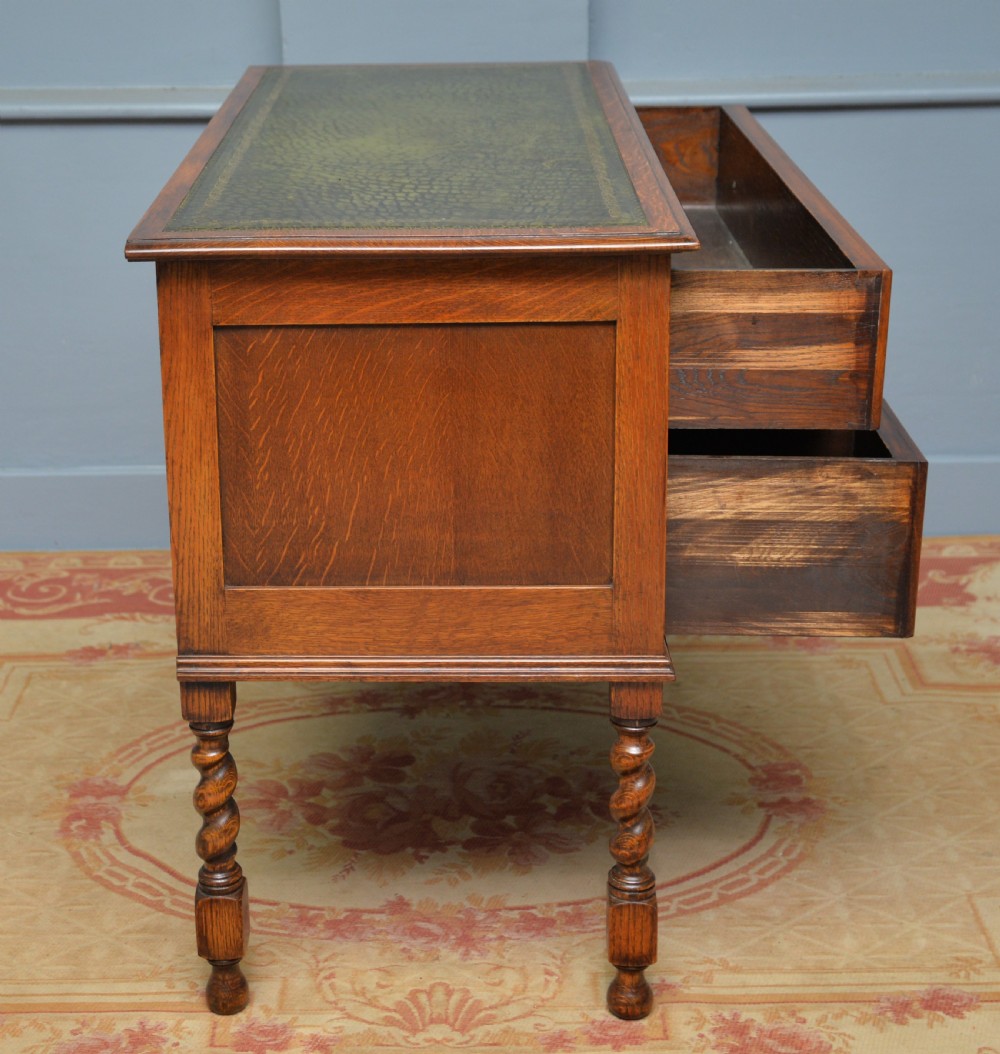 A solid oak writing table featuring a green leather inset top, three drawers with brass drop handles, and turned barley twist legs. View XI