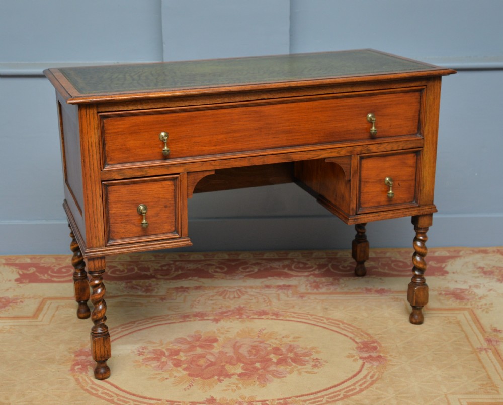 A solid oak writing table featuring a green leather inset top, three drawers with brass drop handles, and turned barley twist legs. View XV