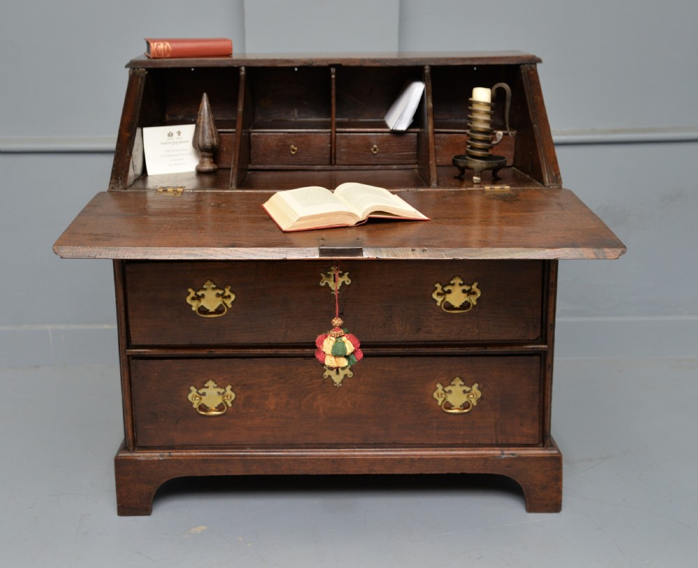 An early 18th-century solid oak writing bureau featuring an angled fall-front that opens to reveal fitted pigeonholes, set over two wide drawers with decorative brass handles.