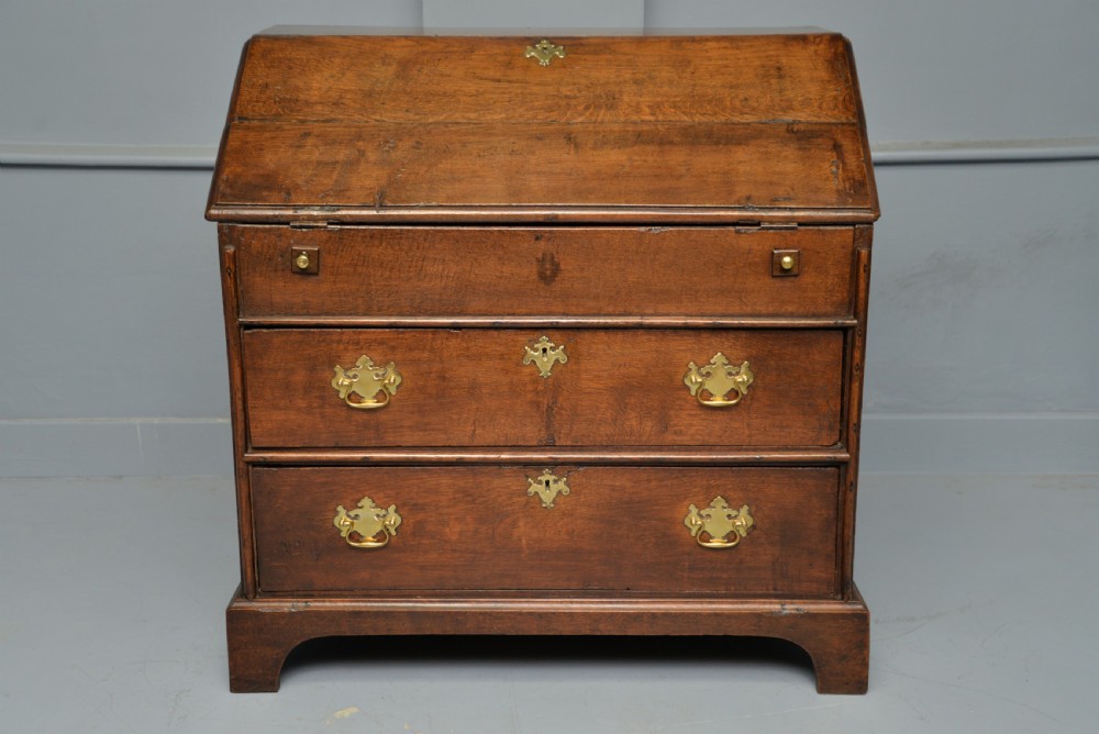 An early 18th-century solid oak writing bureau featuring an angled fall-front that opens to reveal fitted pigeonholes, set over two wide drawers with decorative brass handles. View II