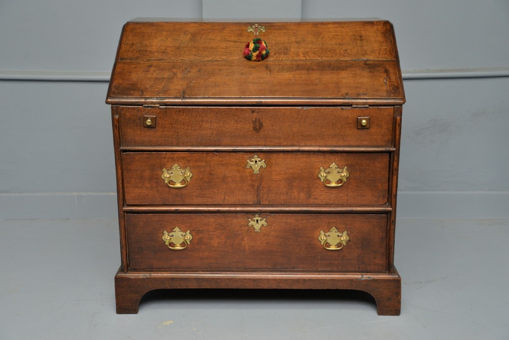 An early 18th-century solid oak writing bureau featuring an angled fall-front that opens to reveal fitted pigeonholes, set over two wide drawers with decorative brass handles. View III