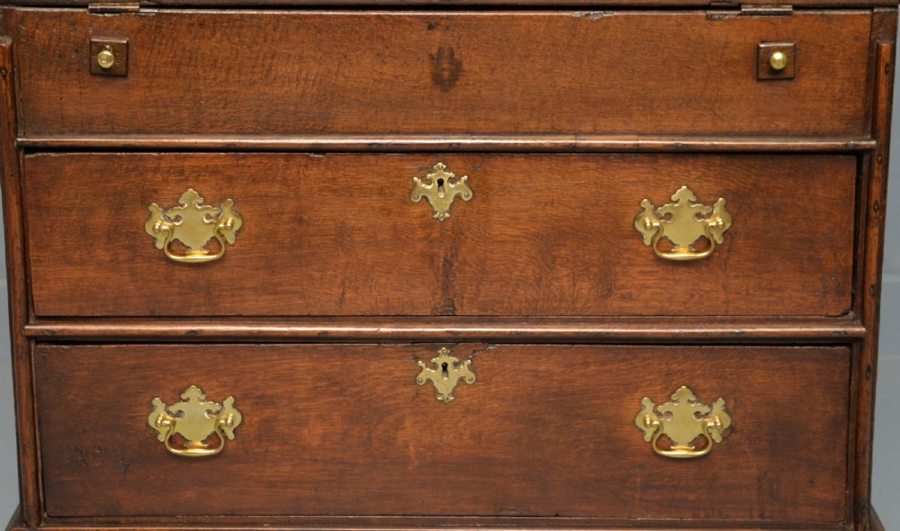 An early 18th-century solid oak writing bureau featuring an angled fall-front that opens to reveal fitted pigeonholes, set over two wide drawers with decorative brass handles. View V