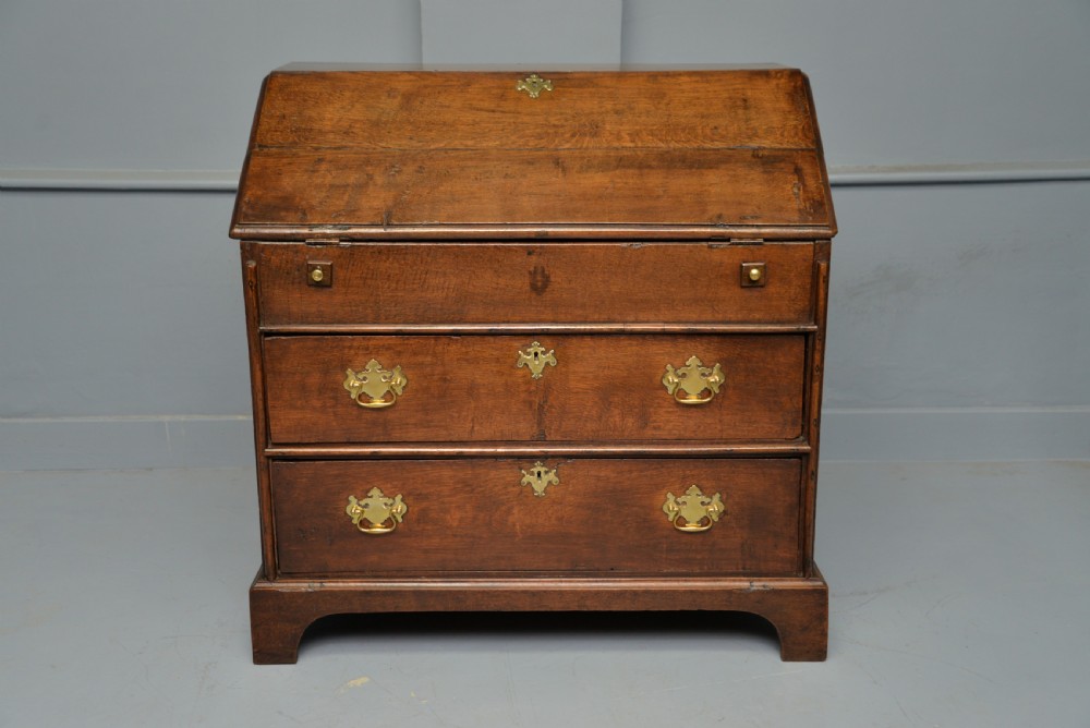 An early 18th-century solid oak writing bureau featuring an angled fall-front that opens to reveal fitted pigeonholes, set over two wide drawers with decorative brass handles. View VI