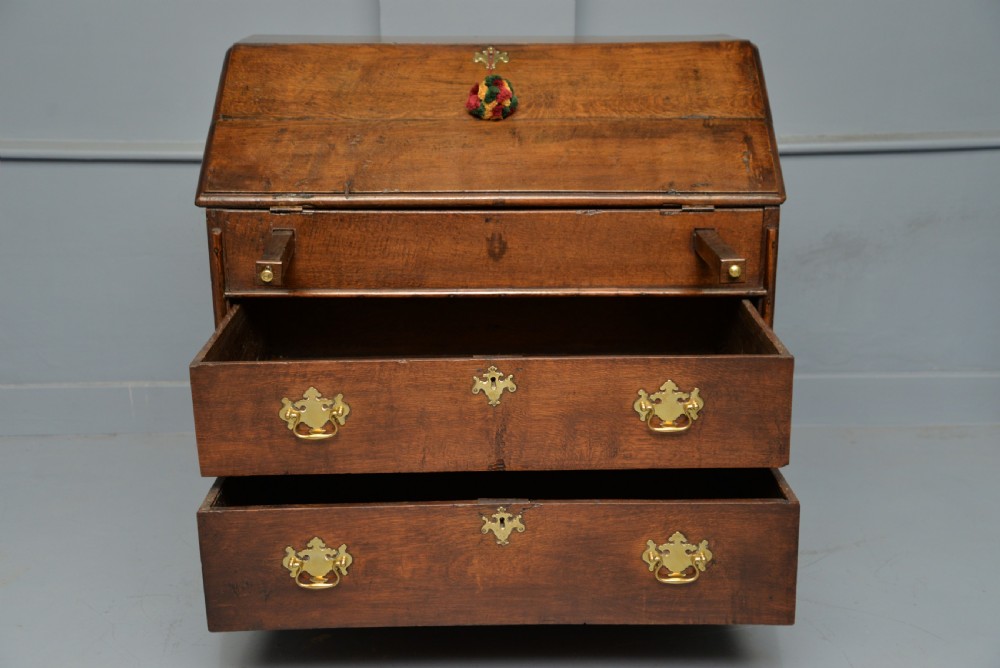 An early 18th-century solid oak writing bureau featuring an angled fall-front that opens to reveal fitted pigeonholes, set over two wide drawers with decorative brass handles. View VII