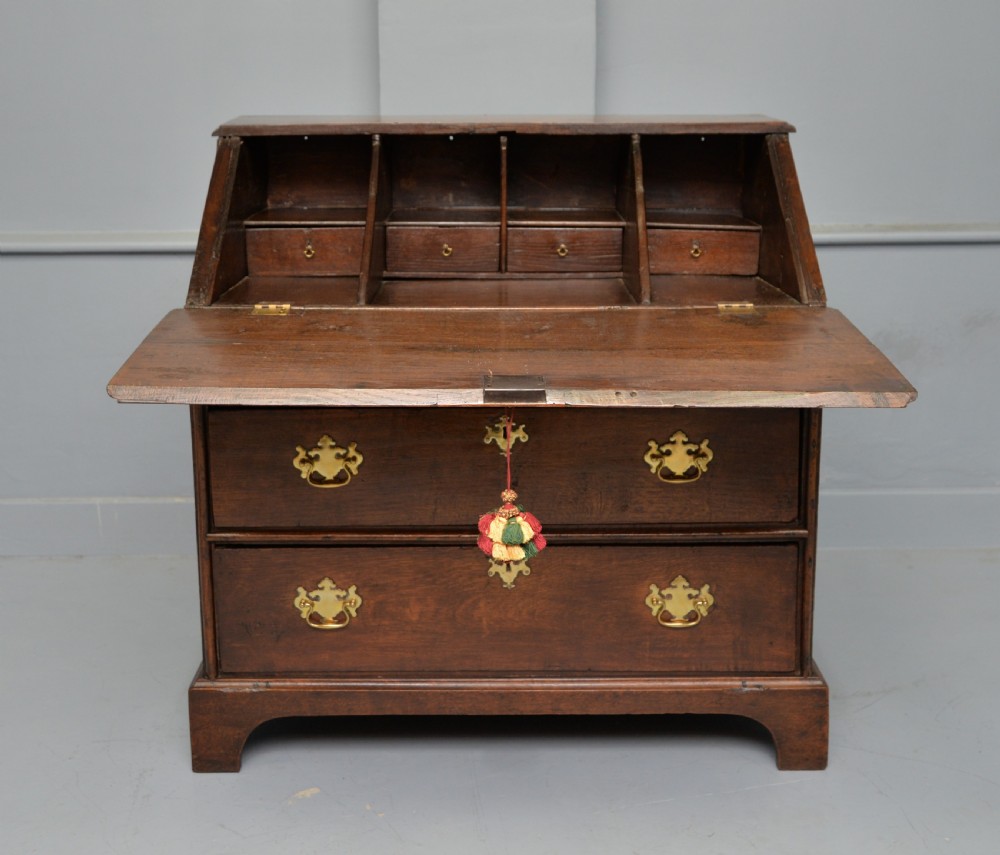 An early 18th-century solid oak writing bureau featuring an angled fall-front that opens to reveal fitted pigeonholes, set over two wide drawers with decorative brass handles. View VIII