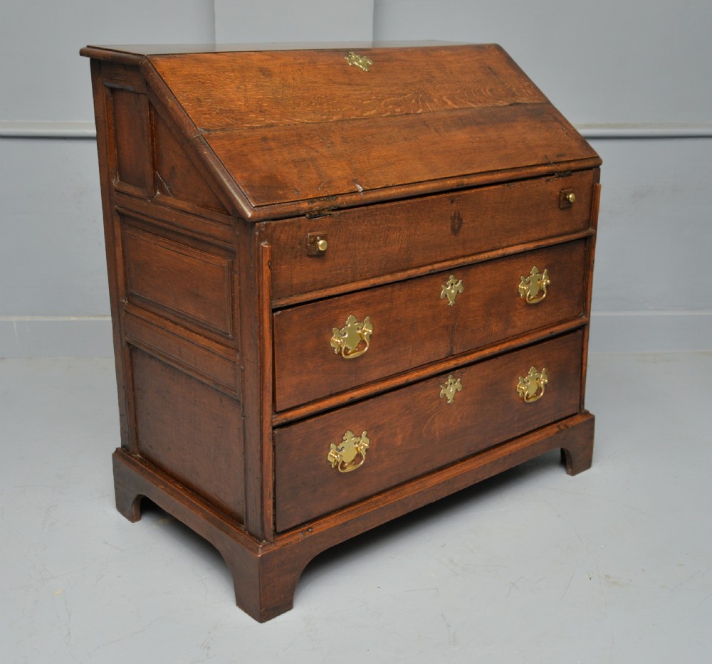 An early 18th-century solid oak writing bureau featuring an angled fall-front that opens to reveal fitted pigeonholes, set over two wide drawers with decorative brass handles. View IX