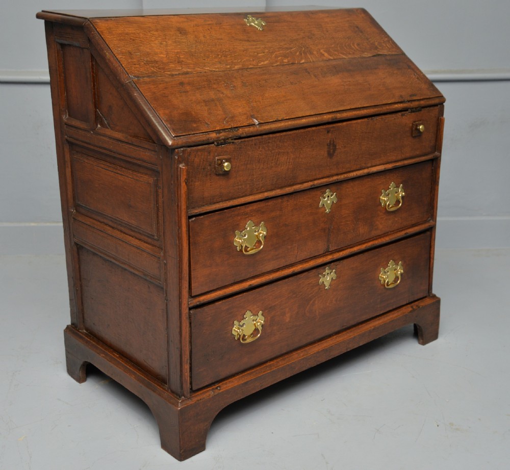 An early 18th-century solid oak writing bureau featuring an angled fall-front that opens to reveal fitted pigeonholes, set over two wide drawers with decorative brass handles. View X