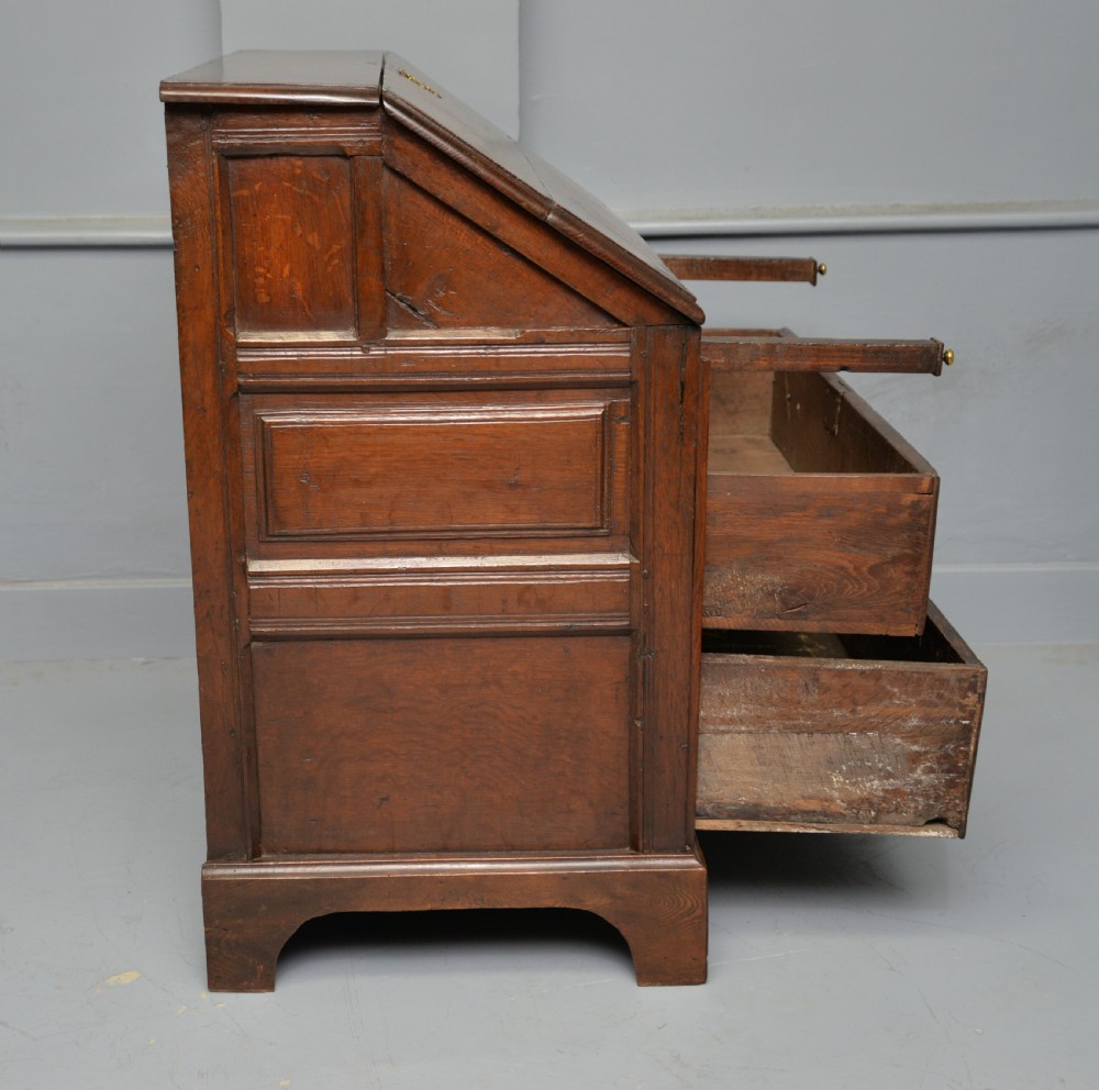 An early 18th-century solid oak writing bureau featuring an angled fall-front that opens to reveal fitted pigeonholes, set over two wide drawers with decorative brass handles. View XI