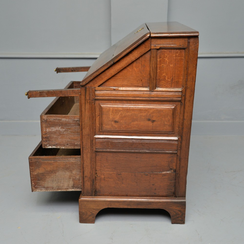 An early 18th-century solid oak writing bureau featuring an angled fall-front that opens to reveal fitted pigeonholes, set over two wide drawers with decorative brass handles. View XIII