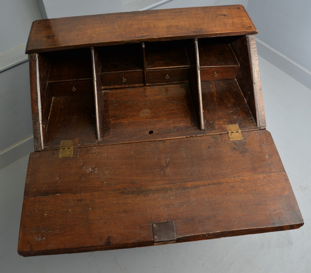 An early 18th-century solid oak writing bureau featuring an angled fall-front that opens to reveal fitted pigeonholes, set over two wide drawers with decorative brass handles. View XIV