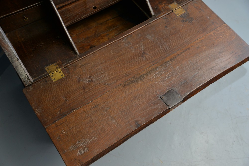 An early 18th-century solid oak writing bureau featuring an angled fall-front that opens to reveal fitted pigeonholes, set over two wide drawers with decorative brass handles. View XVI