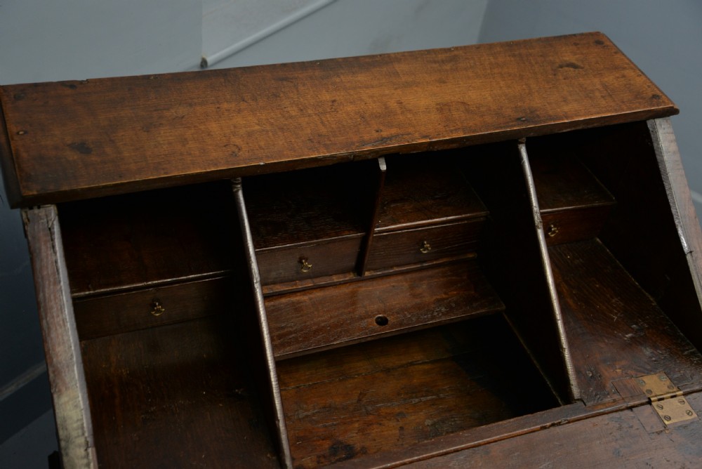 An early 18th-century solid oak writing bureau featuring an angled fall-front that opens to reveal fitted pigeonholes, set over two wide drawers with decorative brass handles. View XVII