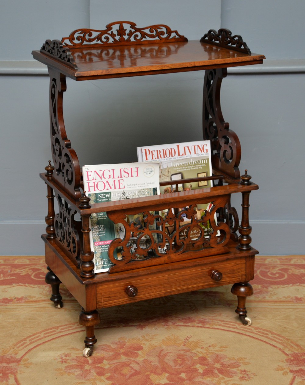 A Victorian burr walnut Canterbury table featuring a top shelf with pierced fretwork, lower magazine compartments, a single drawer, and turned legs with ceramic castors.