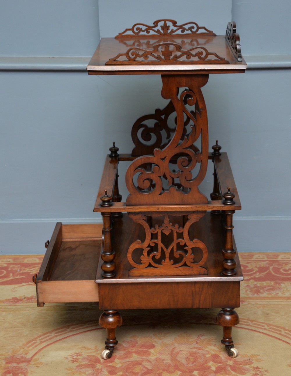 A Victorian burr walnut Canterbury table featuring a top shelf with pierced fretwork, lower magazine compartments, a single drawer, and turned legs with ceramic castors. View X