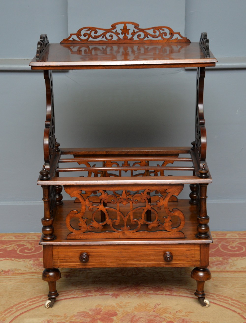 A Victorian burr walnut Canterbury table featuring a top shelf with pierced fretwork, lower magazine compartments, a single drawer, and turned legs with ceramic castors. View XII