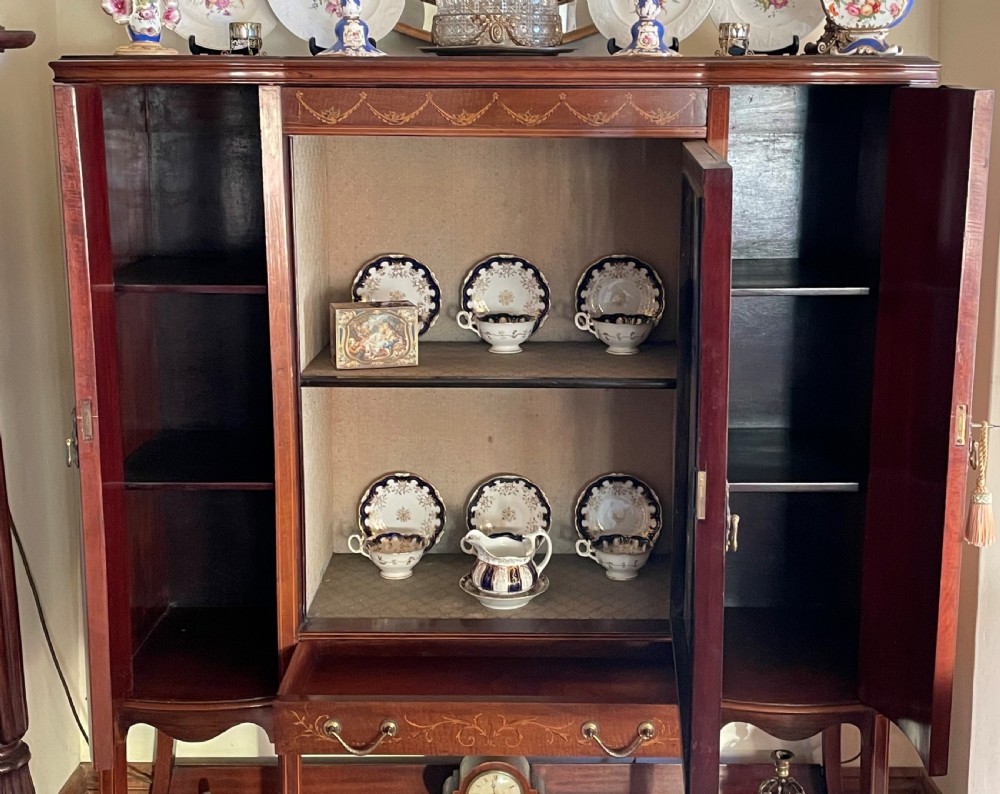 An inlaid mahogany display cabinet featuring a central glazed door with curved astragal glazing bars, flanking curved inlaid cupboard doors, a lower drawer, and an undertier shelf. View VI