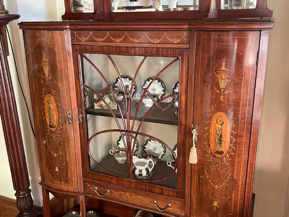 An inlaid mahogany display cabinet featuring a central glazed door with curved astragal glazing bars, flanking curved inlaid cupboard doors, a lower drawer, and an undertier shelf. View XIV
