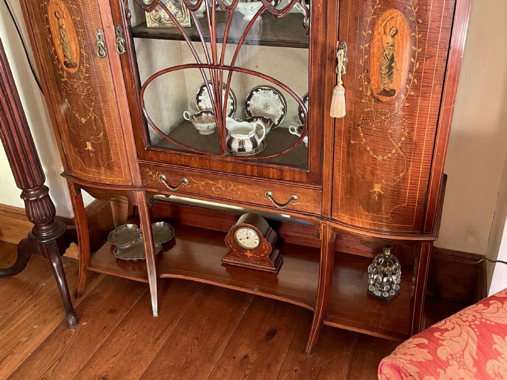 An inlaid mahogany display cabinet featuring a central glazed door with curved astragal glazing bars, flanking curved inlaid cupboard doors, a lower drawer, and an undertier shelf. View XV