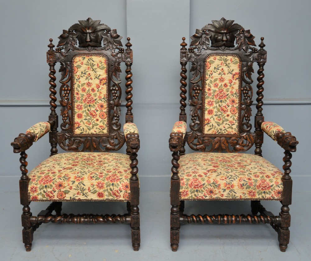 A pair of Victorian dark oak throne chairs featuring Green Man carved crestings, barley twist supports, carved beast armrests, and floral pattern upholstery. View V