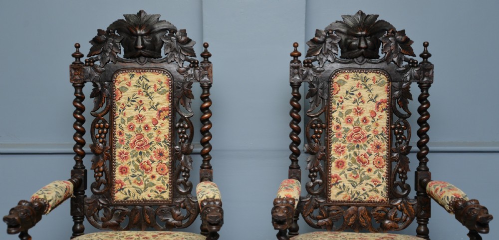A pair of Victorian dark oak throne chairs featuring Green Man carved crestings, barley twist supports, carved beast armrests, and floral pattern upholstery. View VI