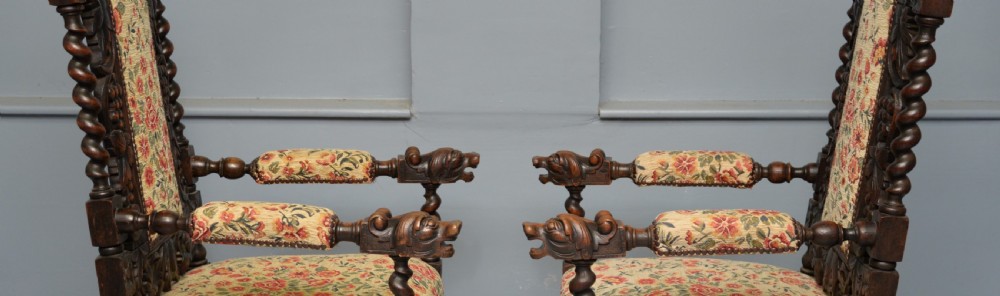 A pair of Victorian dark oak throne chairs featuring Green Man carved crestings, barley twist supports, carved beast armrests, and floral pattern upholstery. View X