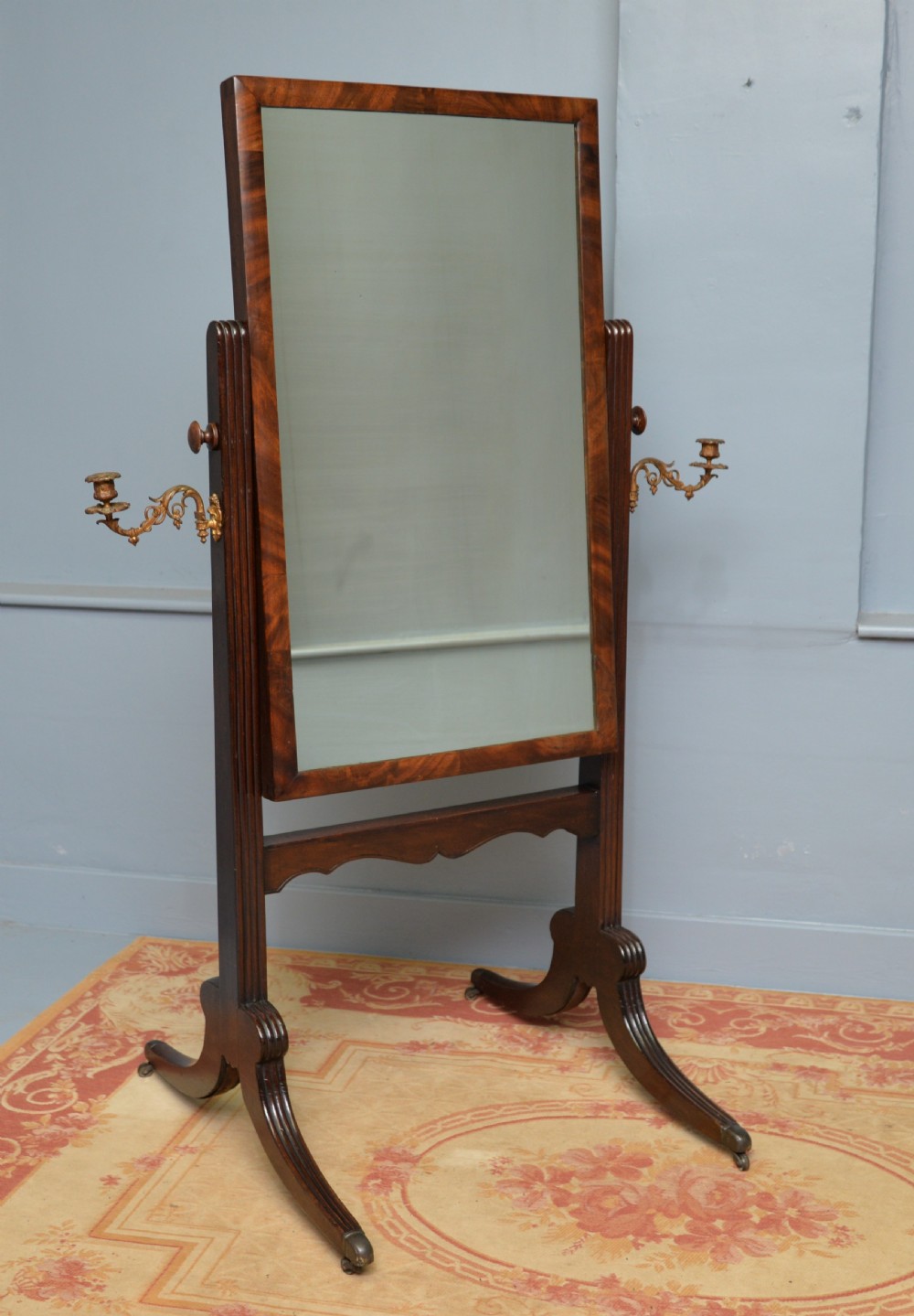 An early 19th-century Regency mahogany cheval mirror featuring a rectangular swivelling glass frame, fluted uprights with twin brass candle sconces, and splayed legs on brass castors.