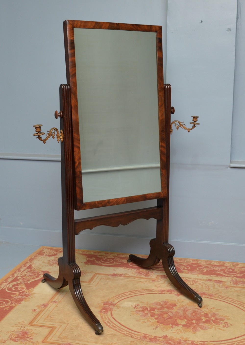 An early 19th-century Regency mahogany cheval mirror featuring a rectangular swivelling glass frame, fluted uprights with twin brass candle sconces, and splayed legs on brass castors. View II