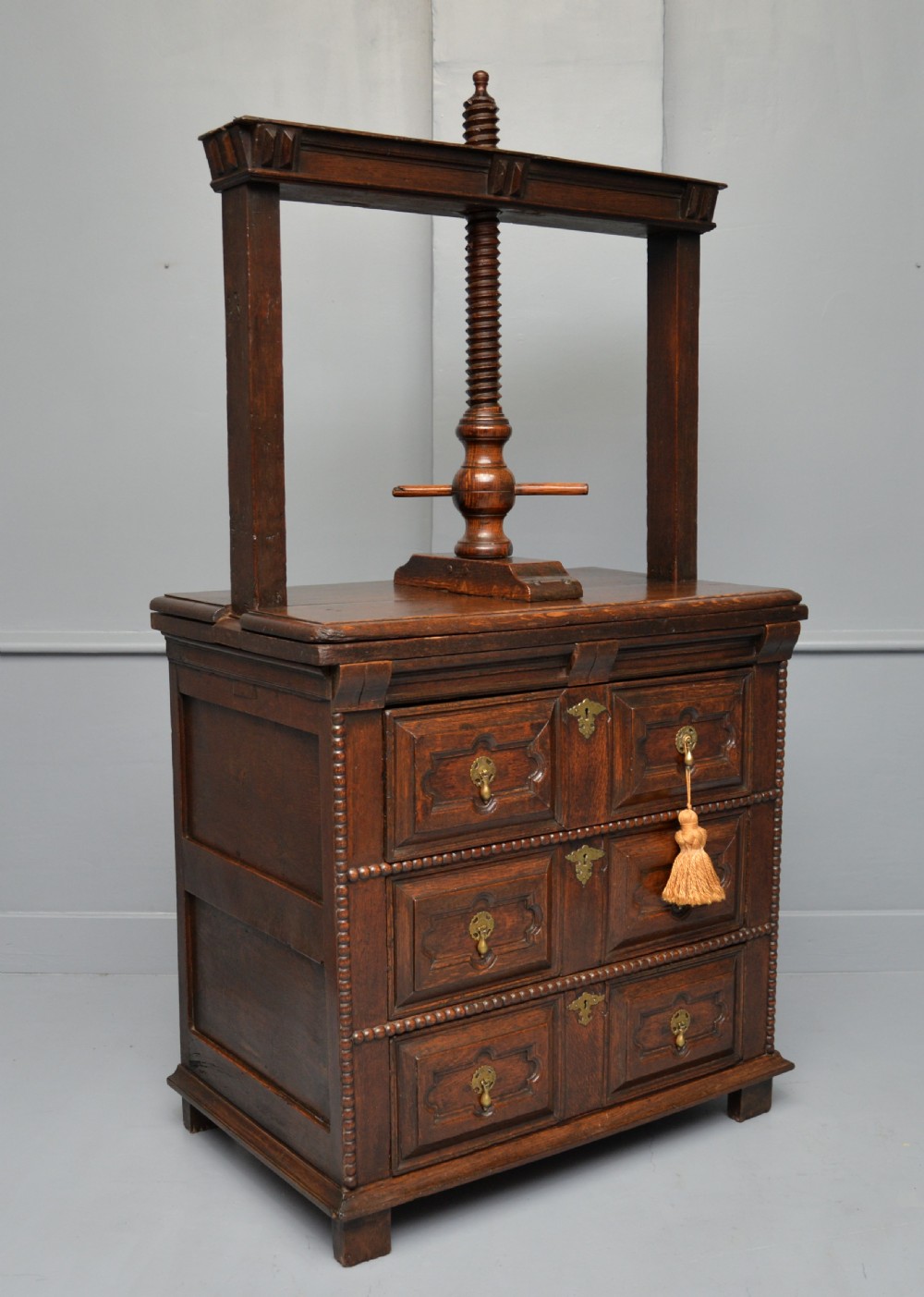 A 17th-century dark oak chest of drawers featuring geometric panelled drawer fronts and an integrated wooden screw linen press mounted on top. View XII