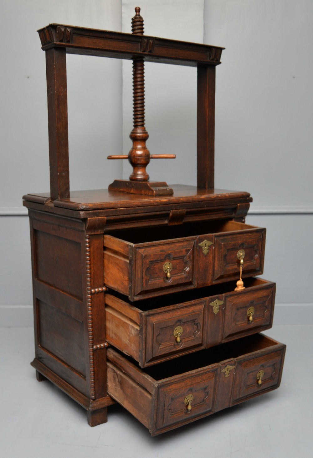 A 17th-century dark oak chest of drawers featuring geometric panelled drawer fronts and an integrated wooden screw linen press mounted on top. View XIII