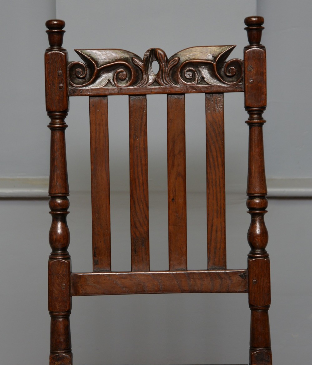 A 17th-century dark oak side chair featuring a carved scroll crest rail, vertical slatted backrest, turned legs, and a solid wood seat. View XVI