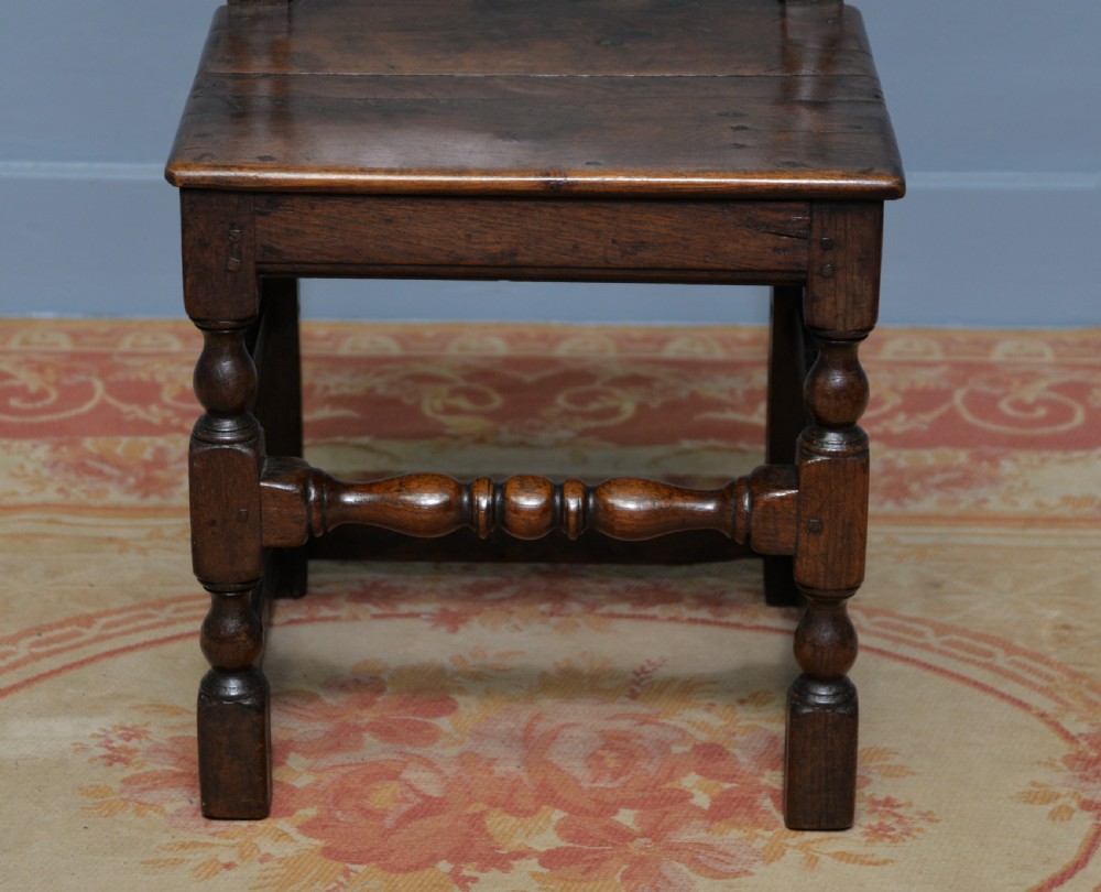A 17th-century dark oak side chair featuring a carved scroll crest rail, vertical slatted backrest, turned legs, and a solid wood seat. View XVIII