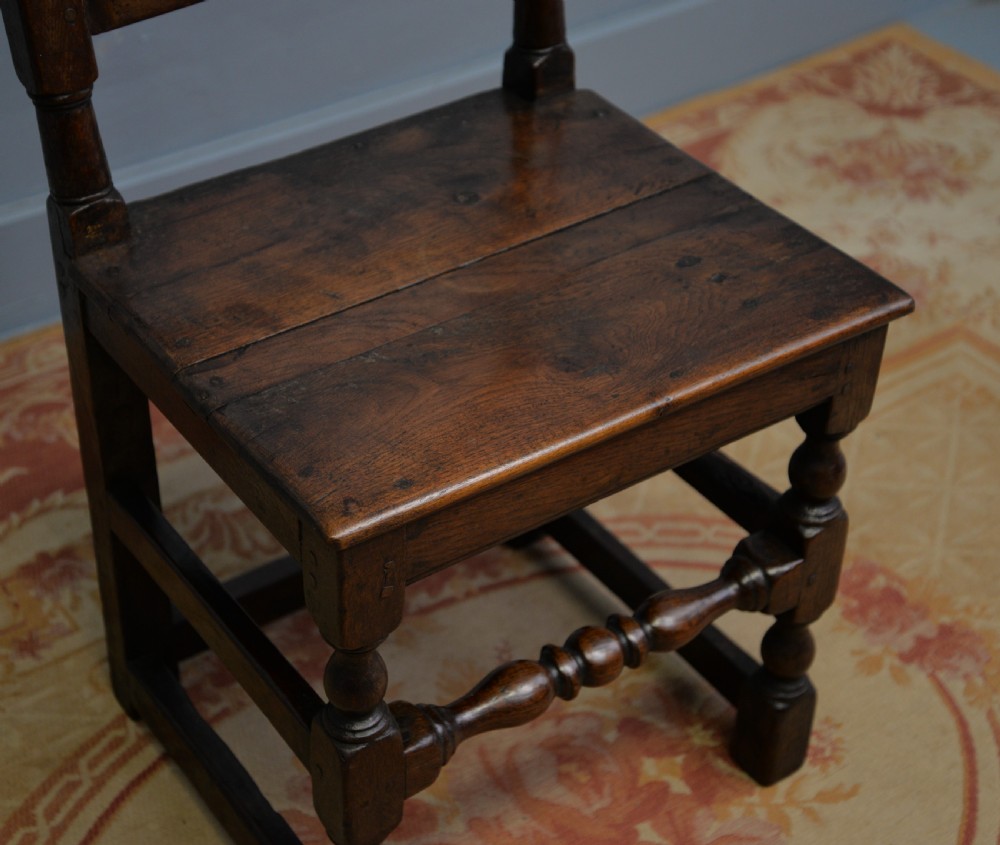 A 17th-century dark oak side chair featuring a carved scroll crest rail, vertical slatted backrest, turned legs, and a solid wood seat. View XX