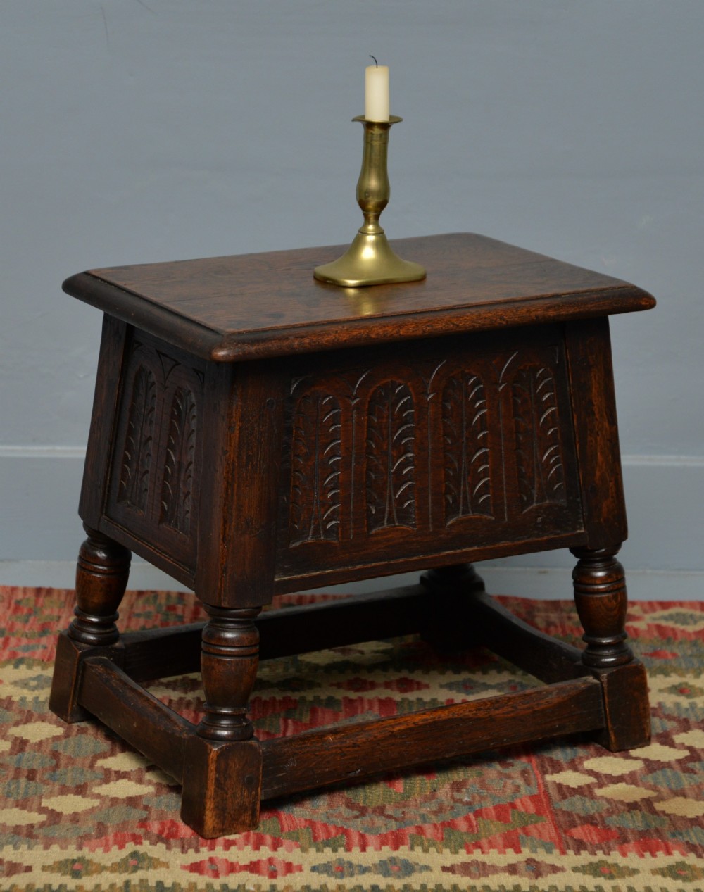 A dark oak box stool featuring carved arched panels, turned baluster legs, and a rectangular top.