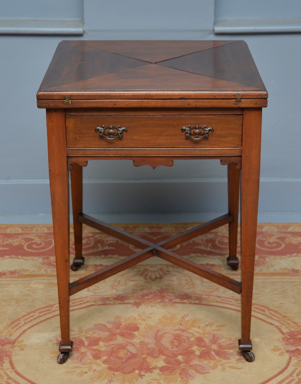 An antique mahogany envelope card table featuring four triangular top flaps, a single drawer with brass drop handles, and tapered legs joined by an X-shaped stretcher.
