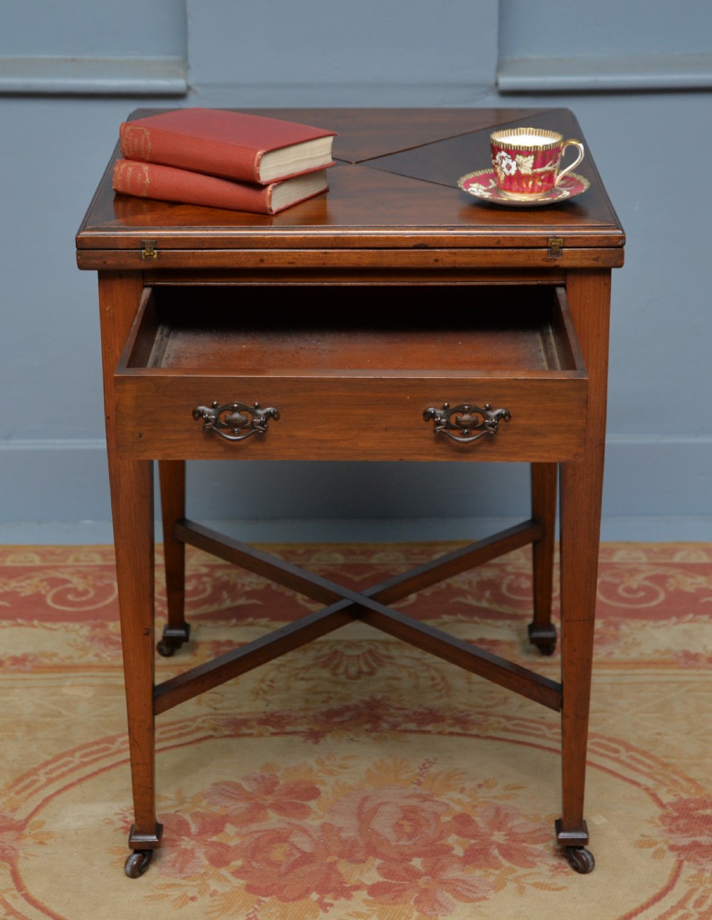 An antique mahogany envelope card table featuring four triangular top flaps, a single drawer with brass drop handles, and tapered legs joined by an X-shaped stretcher. View III