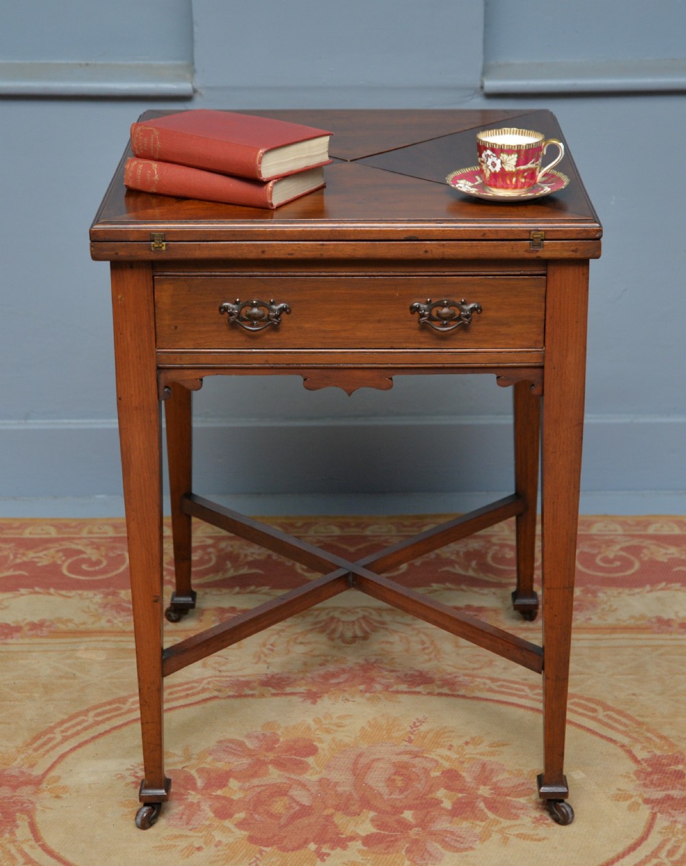 An antique mahogany envelope card table featuring four triangular top flaps, a single drawer with brass drop handles, and tapered legs joined by an X-shaped stretcher. View IV
