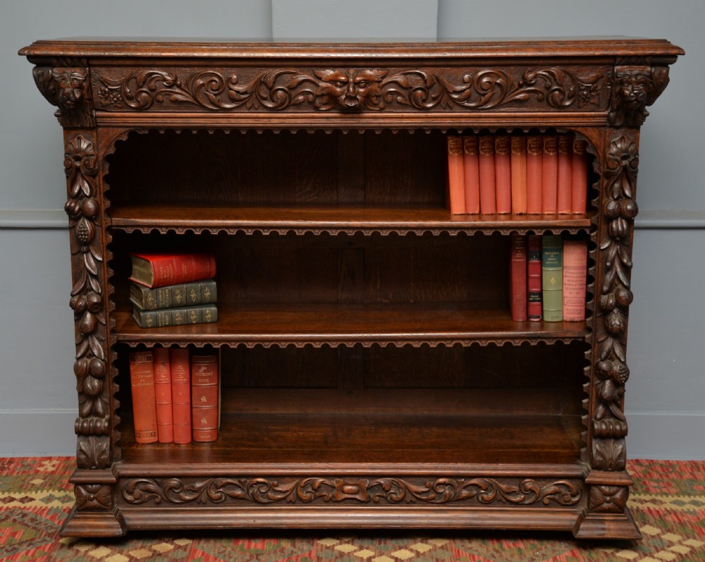 A low solid oak open bookcase decorated with hand-carved Green Man motifs, foliate pilasters, three shelves with scalloped edges, and a plinth base.